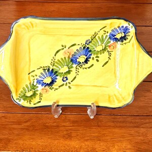 May include: A rectangular yellow ceramic serving tray with scalloped edges and a dark blue rim. The center features a hand-painted diagonal floral pattern consisting of blue and green daisy-like flowers with small orange accents and green leaves.