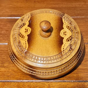 May include: A round wooden lidded box featuring a central knob handle. The lid displays a two-tone finish with light and dark brown sections, accented by intricate laser-cut lace-like patterns and a carved geometric border around the edge.