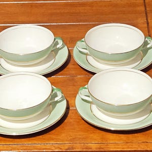 May include: Four matching vintage teacups and saucers. The cups are white with a pale green band and gold trim. The saucers are also pale green with gold trim. The set is arranged on a wooden surface.