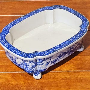 Op de afbeelding: Een ovale keramische plantenbak met een witte binnenkant en een blauw-wit bloemmotief aan de buitenkant. De rand heeft een decoratieve blauwe boord met kleine bloemmotieven. De bak rust op vier kleine, afgeronde voetjes aan de onderzijde.