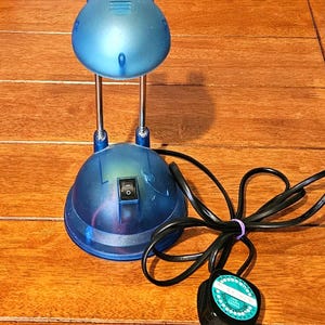 May include: A blue desk lamp with a rounded head and base, supported by two silver metal stems. The lamp has an on/off switch and a black power cord with a plug. The lamp is on a wooden surface.