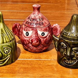 May include: Three ceramic lidded jars with anthropomorphic faces. The left jar is green with the word CUCUMBER. The center jar is reddish-purple with the text PICKLED CABBAGE. The right jar is dark green with the word CUCUMBER and pointed ears.