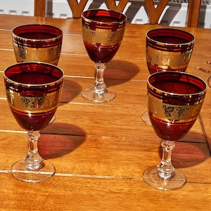 Puede incluir: Cinco copas de vino tinto rubí con ribete dorado y un diseño decorativo de uvas. Las copas están sobre una mesa de madera, mostrando su elegante diseño y estética vintage. Las copas tienen un tallo y base transparentes.