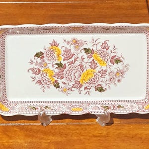 Puede incluir: Bandeja rectangular para servir con un diseño floral. La bandeja presenta un fondo blanco con un borde y un arreglo floral en amarillo, rosa y verde. La bandeja está sostenida por dos soportes de plástico transparente.