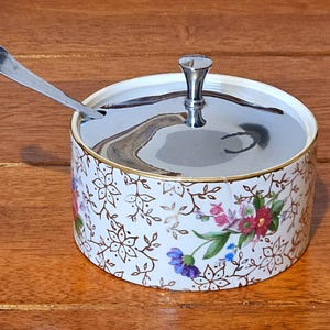 Puede incluir: Un azucarero de cerámica blanca con un diseño floral y dorado, con tapa plateada y una cucharita. El cuenco tiene un borde dorado y está decorado con motivos florales marrones y flores coloridas.