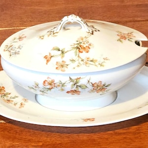 Może przedstawiać: Biała porcelanowa salaterka z pokrywką i pasującym talerzem. Salaterka i pokrywka zdobione wzorem kwiatowym w odcieniach pomarańczu, żółci i zieleni. Uchwyt na pokrywce jest ozdobny.