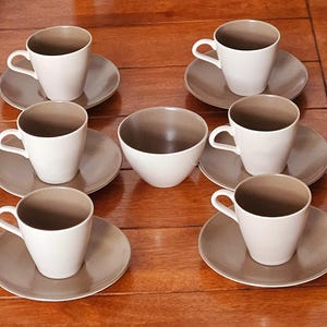 Può includere: Un set di sei tazze da caffè in ceramica bianca con interni marroni e piattini abbinati, insieme a una ciotola in ceramica bianca con interno marrone. Le tazze e i piattini sono disposti su una superficie di legno.