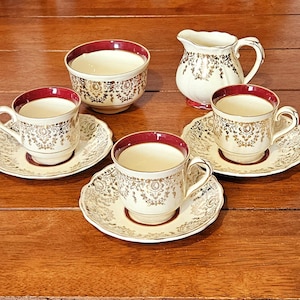 May include: A tea set consisting of three cups with matching saucers, a sugar bowl, and a creamer. Each piece is cream-colored porcelain with a deep red rim and intricate gold filigree patterns. The saucers and cups feature scalloped edges.