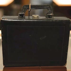 May include: A vintage black briefcase with a leather handle and metal clasp. The rectangular case has a stitched border and shows signs of wear, suggesting age and use. The handle is attached with metal hardware.