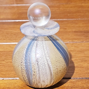Puede incluir: Un frasco de perfume decorativo de vidrio con un tapón redondo y transparente. El frasco tiene forma esférica con una superficie texturizada y moteada y rayas azules y blancas. El frasco está sobre una superficie de madera.