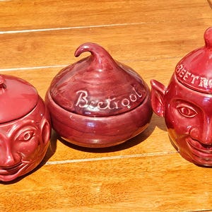 May include: Three dark red ceramic lidded jars. The left jar features a face with a stem-like lid. The center jar is round with the word Beetroot embossed on the lid. The right jar has a face, ears, and a lid with the word Beetroot embossed.