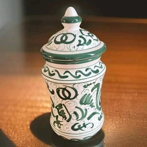 Puede incluir: Un tarro de cerámica blanco con tapa verde y diseños florales verdes. El tarro tiene un cuerpo redondeado y un cuello estrecho.