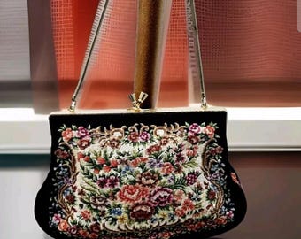 Pode incluir: Bolsa vintage preta com um design de tapeçaria floral. A bolsa apresenta um padrão bordado colorido de flores e folhagem em um fundo creme. Possui uma moldura preta, fecho de pressão e alça de corrente prateada.
