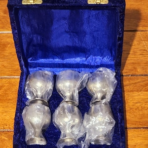 May include: A set of six silver-toned goblets, each individually wrapped in clear plastic, are nestled inside a deep blue velvet-lined box. The box has a hinged lid with gold-colored clasps, set against a wooden surface.