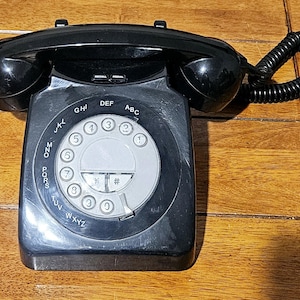 Op de afbeelding: Een zwarte draaischijftelefoon met een handset en een opgerold snoer. De telefoon heeft een ronde draaischijf met cijfers en letters, waaronder de letters ABC, DEF, GHI, JKL, MNO, PQRS, TUV en WXYZ. De telefoon staat op een houten oppervlak.