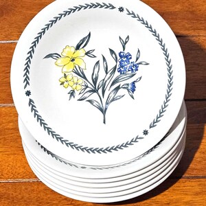 Puede incluir: Una pila de platos de cerámica blancos con un diseño floral. Cada plato presenta un ramo central de flores amarillas y azules con hojas verdes. Un borde de hojas grises y pequeñas estrellas rodea el borde.