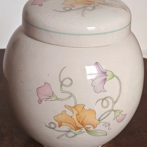 May include: A white ceramic jar with a lid, featuring a floral design of purple, pink, yellow, and green sweet peas. The jar has a crackled glaze finish.