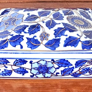 May include: Rectangular porcelain trinket box with a white base and intricate blue floral and leaf designs. The lid and sides feature detailed hand-painted flowers and foliage, accented with gold trim. A decorative storage container.