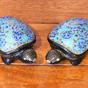 Könnte beinhalten: Zwei dekorative Schildkrötenfiguren. Die Schildkrötenpanzer sind mit einem Blumenmuster in Blau- und Türkistönen verziert, das mit Gold umrandet ist. Die Schildkröten haben schwarze Körper und Köpfe mit weißen Punkten.
