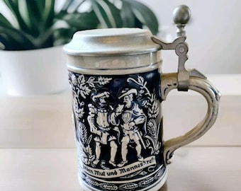 May include: A blue and white ceramic beer stein with a pewter lid and handle. The stein features a scene of two men in traditional German clothing with the text "Ehren Mut und Manneskraft".