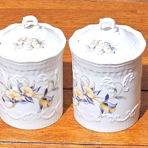 May include: Two white ceramic canisters with lids. Each canister features a floral design with yellow and blue flowers and decorative scrollwork. The lids have a small loop handle.