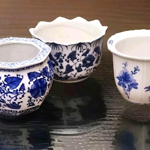 以下が含まれることがあります： 青い花柄の白い陶器の植木鉢が3つ。植木鉢は、六角形、スカラップエッジ、丸みを帯びた形状など、さまざまな形をしています。青い花柄は植木鉢によって異なります。