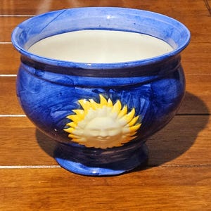 Op de afbeelding: Blauwe keramische bloempot met een witte binnenkant en een decoratief zonontwerp. De pot heeft een ronde vorm met een uitlopende rand en een voetstuk. Het zonontwerp heeft een lachend gezicht met gele stralen.