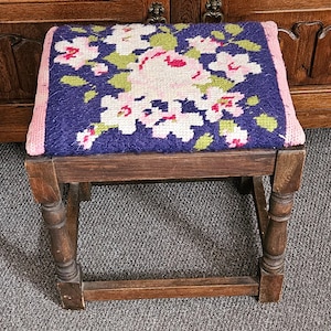 Puede incluir: Taburete pequeño de madera con asiento de tapiz. El asiento presenta un diseño floral en blanco, rosa y verde sobre un fondo azul oscuro, enmarcado por un borde rosa. Las patas son de color marrón oscuro y torneadas.