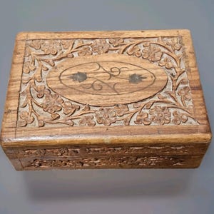May include: A rectangular wooden box with intricate floral carvings. The box features a central oval design with a floral and leaf pattern. The exterior is adorned with detailed carvings of flowers and foliage, showcasing a natural wood finish.