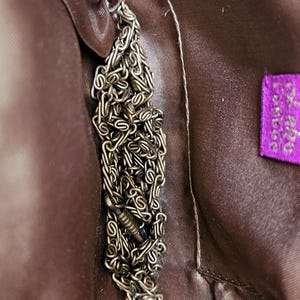 May include: Close-up of a brown handbag interior with a silver chain and a purple label. The label has white text. The handbag's interior is a smooth, satiny material.