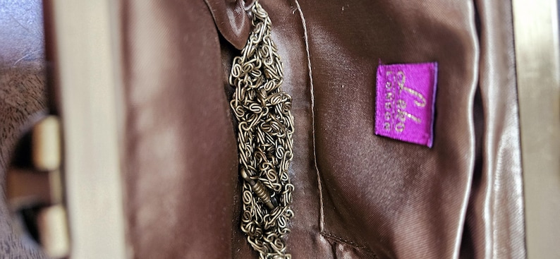 May include: Close-up of a brown handbag interior with a silver chain and a purple label. The label has white text. The handbag's interior is a smooth, satiny material.