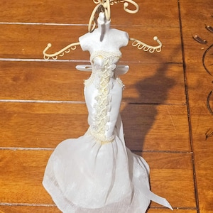 May include: A decorative white dress form with a long, flowing skirt. The form features a detailed bodice with lace accents and a decorative headpiece. The dress form is designed to hold jewelry or small accessories.