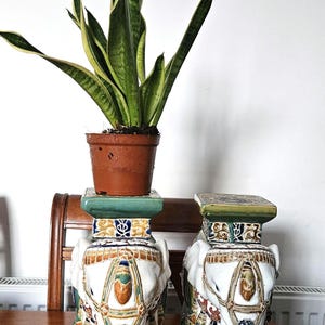 May include: Two white ceramic elephant garden stools with colourful detailing. One stool supports a potted plant with green and yellow leaves. The stools are on a wooden table.