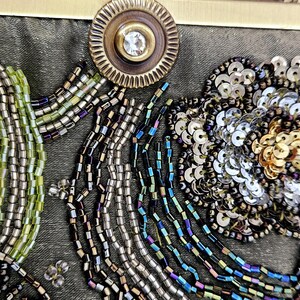 May include: Close-up of an olive green beaded clutch bag. The bag features a floral design with silver sequins and gold beads. A cactus-like design is also present, along with a decorative clasp.