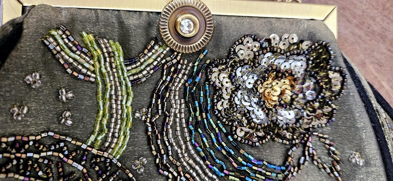 May include: Close-up of an olive green beaded clutch bag. The bag features a floral design with silver sequins and gold beads. A cactus-like design is also present, along with a decorative clasp.