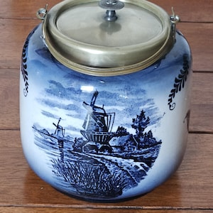 May include: A blue and white ceramic jar with a metal lid and handle. The jar features a landscape scene with windmills and a village. The lid has a small decorative knob. The jar sits on a wooden surface.