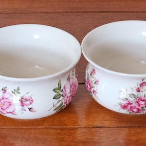 Könnte beinhalten: Zwei weiße Keramikschalen mit einem floralen Rosendesign. Die Schalen haben eine breite, flache Form und sind mit rosa Rosen und grünen Blättern verziert. Die Schalen stehen auf einer Holzoberfläche.