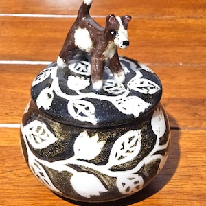 Puede incluir: Un tarro de cerámica con tapa, con una figura de perro marrón y blanco en la parte superior. El tarro es negro con diseños de hojas blancas y detalles dorados. El tarro está sobre una superficie de madera.