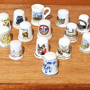 May include: A selection of ceramic thimbles, each with a distinct design. The thimbles showcase various images, including landmarks, landscapes, and patterns. The thimbles are white with gold trim and are arranged on a wooden surface.