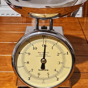 May include: A vintage chrome kitchen scale with a large, round dial and a stainless steel bowl. The dial has black numbers on a cream background and reads "SALTER". The scale measures up to 5 kg.