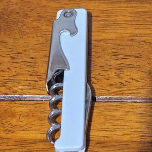 Pode incluir: Um saca-rolhas branco e prateado com um abridor de garrafas embutido. O saca-rolhas tem um parafuso prateado e uma alavanca articulada para facilitar a remoção da rolha. A pega é de cor azul clara. A ferramenta foi concebida para abrir garrafas de vinho.