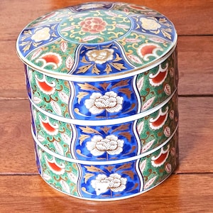 May include: A three-tiered, round ceramic container with a lid. The container features a vibrant design with blue, green, red, and gold floral patterns. White floral designs are centered on each tier. The container is likely used for storage or display.
