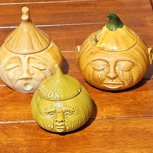 May include: Three ceramic onion-shaped jars with lids, each featuring a sculpted face with tear-like markings. The two larger jars are yellow-brown, while the smaller one is olive green. The word ONION is embossed on the lids of the jars.
