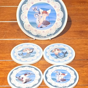 May include: A set of four round coasters and a matching plate. Each coaster and the plate feature a whimsical illustration of a mermaid, cherub, and whale in a blue and white seashell design. The plate is elevated on a white stand.