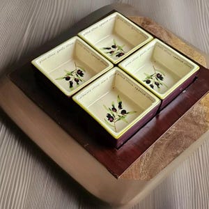 Puede incluir: Un juego de cuatro cuencos cuadrados de cerámica para aceitunas con un diseño de rama de olivo pintada. Los cuencos son de color crema con un borde morado oscuro y se colocan sobre una bandeja de madera. El texto en los cuencos indica los tipos de aceitunas: Kalamata, verdes, deshuesadas y españolas.