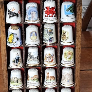 May include: A collection of decorative thimbles displayed in a wooden case. Each white thimble features a unique design, including images of landmarks, animals, and text. The case has a dark brown finish and a metal handle.