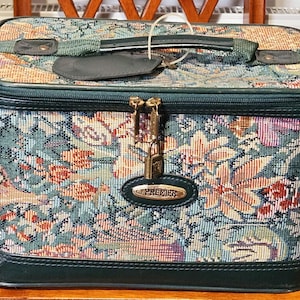 May include: A rectangular vanity case featuring a multi-colored floral tapestry pattern with dark green trim. The case includes a top handle, a luggage tag, a double zipper closure with a small brass padlock, and a gold-tone plaque labeled PREMIER.