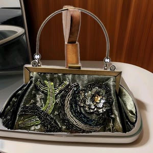 May include: An olive green satin handbag with a decorative floral design. The bag features a metal frame and handle, with beaded and sequined embellishments in black, green, and silver. The bag measures approximately 25 cm wide.