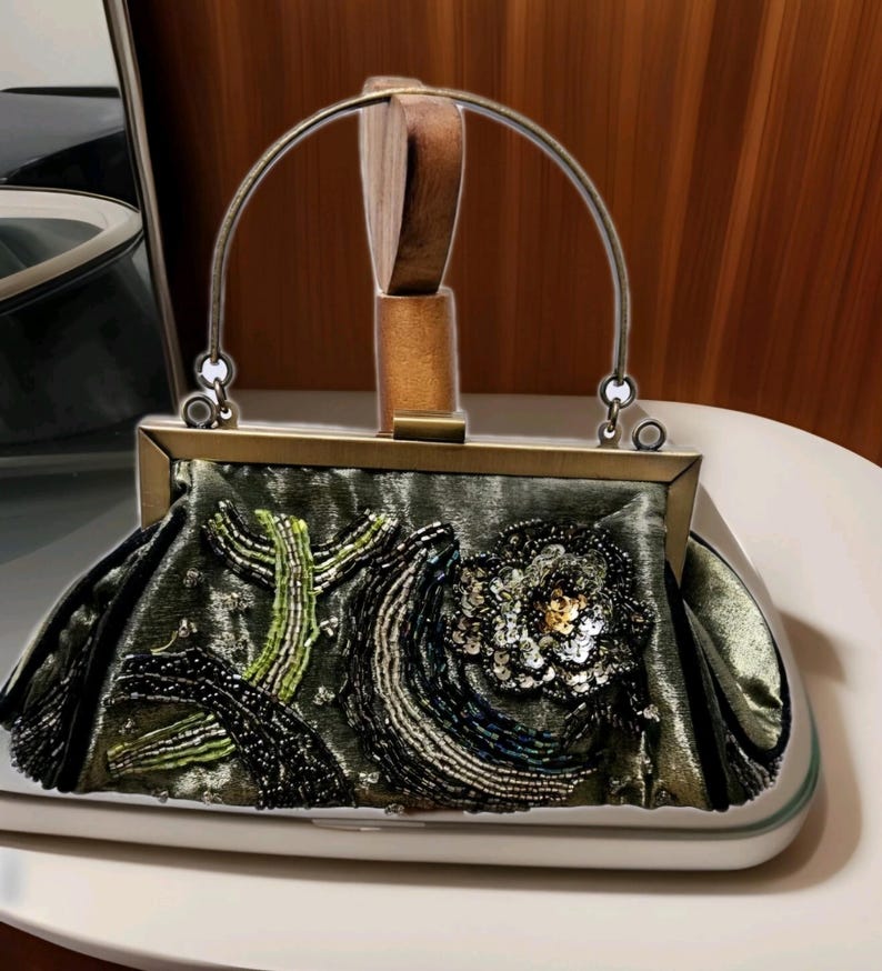 May include: An olive green satin handbag with a decorative floral design. The bag features a metal frame and handle, with beaded and sequined embellishments in black, green, and silver. The bag measures approximately 25 cm wide.