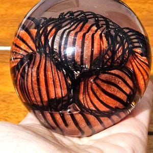 May include: A clear glass orb with a dark, smoky top. Inside, there are several orange and black striped orbs, creating a unique decorative piece. The orb is held in a hand, showcasing its size and design.